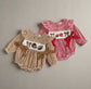 Two baby rompers, one brown checkered and one red checkered with decorative elements, on a beige background.